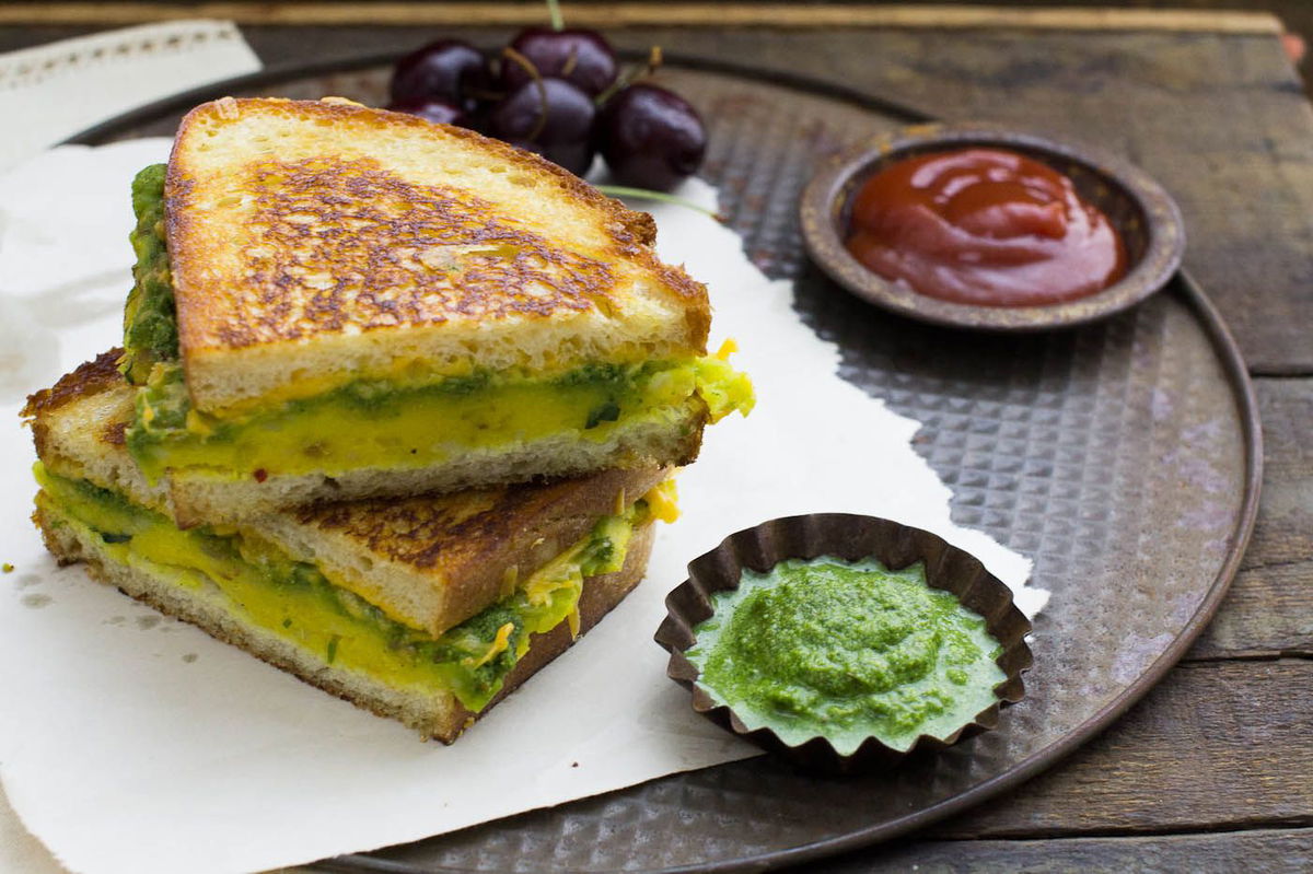masala cheese toast sandwich halves layered one above another with cherries, ketchup and chutney on white paper and round tray